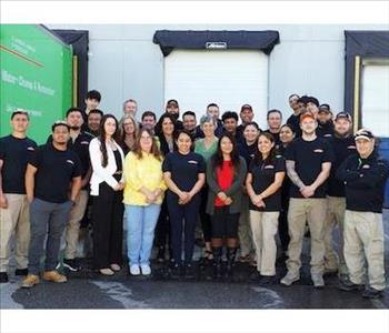 The SERVPRO of Gaithersburg/Germantown Team, in SERVPRO uniforms, standing in 3 rows facing front, with SERVPRO trucks behind